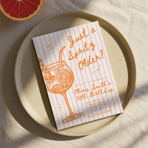May include: A birthday invitation with the text "Just a Spritz Older!" in orange script. The invitation features a cocktail illustration and pink striped background. The invitation is on a beige plate with a grapefruit half in the corner.