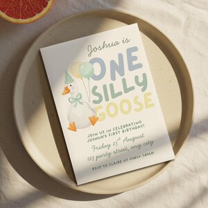 May include: A birthday invitation with the text "Joshua is ONE SILLY GOOSE" in blue, green, and yellow. The invitation features a cartoon goose holding balloons. The invitation is on a beige plate with a slice of orange.