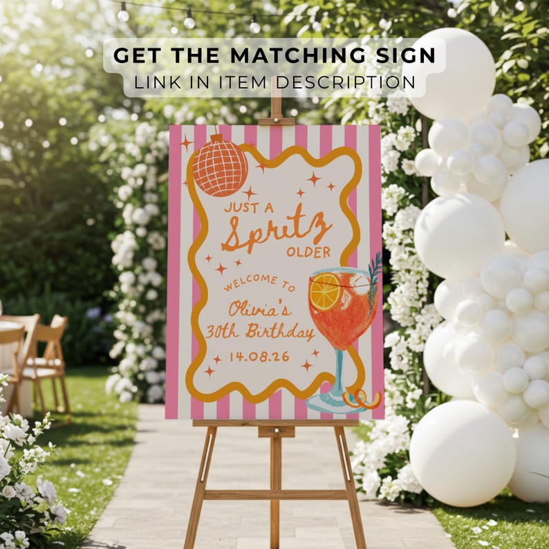 Op de afbeelding: Een verjaardagsfeestbord op een houten ezel. Het bord heeft de tekst "JUST A Spritz OLDER" en "WELCOME TO Olivia's 30th Birthday 14.08.26". Het heeft een cocktail illustratie en een roze en witte gestreepte achtergrond. Buiten.