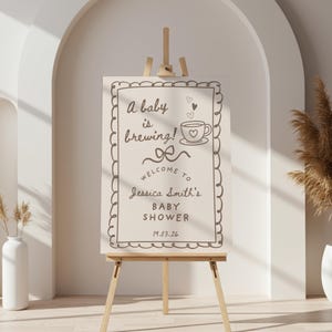 May include: A baby shower welcome sign on a wooden easel. The sign has a decorative border and reads "A baby is brewing!" with a coffee cup illustration. The sign welcomes guests to Jessica Smith's baby shower.