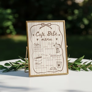 以下が含まれることがあります： 金色のフレームに入ったカフェメニュー。メニューには「Cafe Bébé menu」の文字と、エスプレッソ、ラテ、カプチーノなどのコーヒーメニューが記載されています。その他のオプションには、オーツミルクとバニラが含まれています。