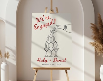 Puede incluir: Un letrero rectangular sobre un caballete de madera con el texto rojo We're Engaged! sobre un dibujo lineal negro de una botella de champán vertiendo en una torre de seis copas, con el texto rojo Ruby + Daniel y 30 de noviembre de 2026.