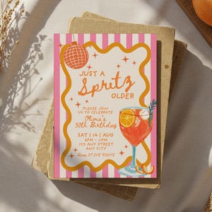 Op de afbeelding: Een uitnodiging voor een feestje met een roze en witte gestreepte achtergrond en een gele rand. De uitnodiging luidt "Just a Spritz Older" en bevat details voor Olivia's 30e verjaardag, met een cocktailillustratie en discobal.