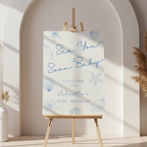 Puede incluir: Un letrero de bienvenida para baby shower sobre un caballete de madera. El letrero es de color crema con ilustraciones de conchas marinas azules y el texto "Sea You Soon Baby. Welcome to Juliette's Baby Shower."