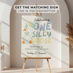 May include: A birthday sign on an easel features a cartoon goose holding balloons. The sign reads "Celebrating One Silly Goose" and welcomes guests to Joshua's first birthday on August 23rd. The sign is light-colored with pastel accents.
