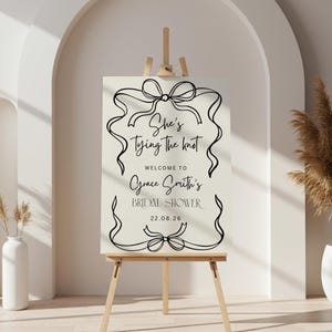 Puede incluir: Un letrero color crema sobre un caballete de madera con texto negro que dice "She's Tying the Knot" y "Welcome to Grace Smith's Bridal Shower 22.08.26". El letrero está decorado con diseños de cinta.
