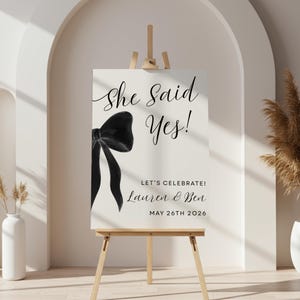 May include: A wedding sign on a wooden easel. The sign reads "She Said Yes!" in elegant script, with a black bow illustration. Below, it says "LET'S CELEBRATE!" followed by names and the date, May 26th, 2026.
