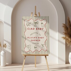 May include: A baby shower sign on a wooden easel. The sign has a cream background with vertical stripes and a red wavy border. The text "CIAO BABY" and "WELCOME TO OLIVIA'S BABY SHOWER" are displayed, along with baby-themed illustrations.