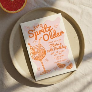May include: A birthday invitation with the text "just a Spritz Older" in orange. The invitation includes a graphic of a cocktail and the details for Olivia's 30th birthday. The invitation is on a beige plate with a grapefruit slice.