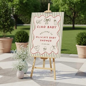 May include: A wooden easel displays a baby shower sign with the text "CIAO BABY WELCOME TO OLIVIA'S BABY SHOWER 17.02.26". The sign has a cream background with green stripes and red accents. Decorative elements include a bow, bottle, and baby clothes.