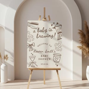 Puede incluir: Un letrero de baby shower de color beige claro sobre un caballete de madera. El letrero presenta ilustraciones con temática de café y el texto "a baby is brewing!" junto con el nombre "Lauren Smith's" y la fecha 07.10.26.