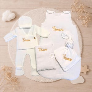 Peut inclure: Ensemble de vêtements et accessoires blancs pour bébé, comprenant un sac de couchage, un bonnet, une couverture et une tenue. Chaque article est brodé du nom "Eleonore" en fil doré. L'ensemble est disposé sur un tapis crème.
