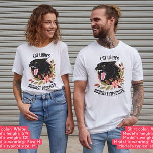White anti-Trumpism t-shirt featuring the words "Cat Ladies Against Fascists" surrounding an illustration of a fierce black cat with florals in hues of rust orange, eggplant, and olive green.