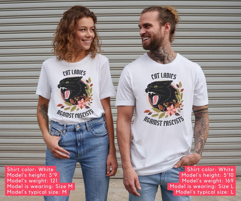 White anti-Trumpism t-shirt featuring the words "Cat Ladies Against Fascists" surrounding an illustration of a fierce black cat with florals in hues of rust orange, eggplant, and olive green.