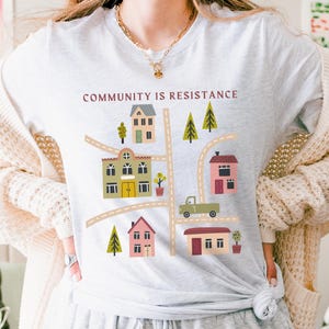 May include: A light gray t-shirt with a graphic design of a neighborhood with houses and trees. The text "COMMUNITY IS RESISTANCE" is printed in maroon on the shirt.