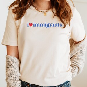 May include: Cream-colored t-shirt with the text "i ❤️ immigrants" in blue and red. The shirt has short sleeves and a crew neck. A light-colored sweater is draped over the shoulders.