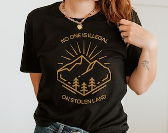 May include: Black t-shirt with a gold graphic design of a mountain range with trees and the text "No one is illegal on stolen land."
