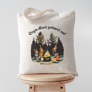 May include: A white tote bag with the text "Cops don't protect us!" printed in black. The bag features a colorful illustration of four animals gathered around a campfire in a snowy forest.