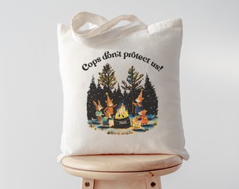 May include: A white tote bag with the text "Cops don't protect us!" printed in black. The bag features a colorful illustration of four animals gathered around a campfire in a snowy forest.
