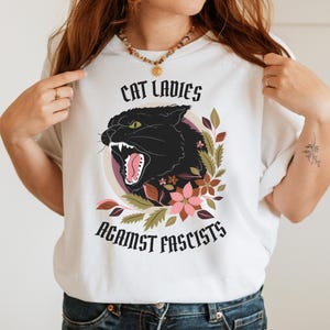 White anti-Trumpism t-shirt featuring the words "Cat Ladies Against Fascists" surrounding an illustration of a fierce black cat with florals in hues of rust orange, eggplant, and olive green.