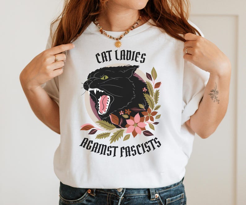 White anti-Trumpism t-shirt featuring the words "Cat Ladies Against Fascists" surrounding an illustration of a fierce black cat with florals in hues of rust orange, eggplant, and olive green.