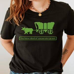 May include: Black t-shirt with a green pixelated image of a wagon pulled by an ox. The text "You have died of corporate greed." is printed below the image.