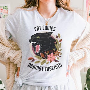Ash anti-Trumpism t-shirt featuring the words "Cat Ladies Against Fascists" surrounding an illustration of a fierce black cat with florals in hues of rust orange, eggplant, and olive green.