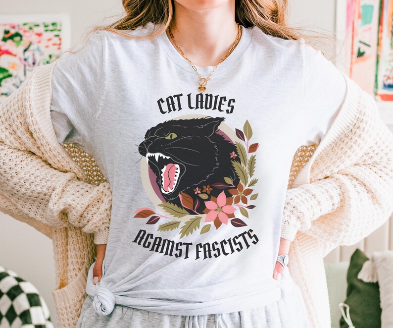 Ash anti-Trumpism t-shirt featuring the words "Cat Ladies Against Fascists" surrounding an illustration of a fierce black cat with florals in hues of rust orange, eggplant, and olive green.