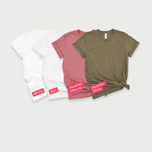 May include: Four t-shirts in different colors: white, ash, heather mauve, and heather olive. The shirts are folded and laid out on a white background.