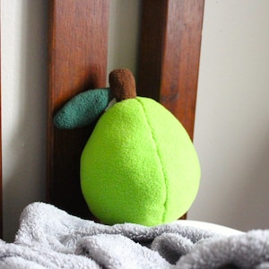 May include: A plush, lime green pear-shaped toy with a brown stem and a green leaf. The soft toy is resting on a gray blanket, with a wooden bed frame in the background.
