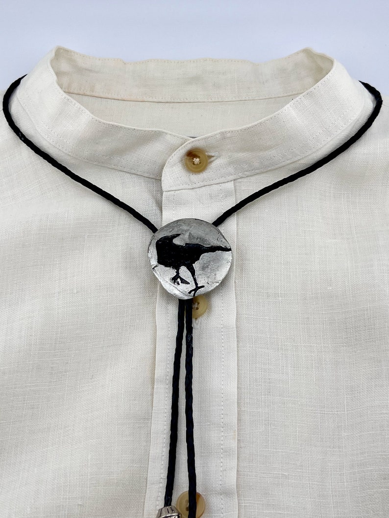 Handmade bolo tie black crow on pewter showing leather braided black cord. Crow bolo tie draped on off-white linen blouse.