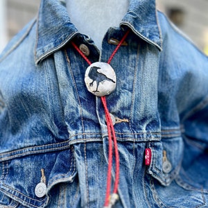 Handmade Black Crow Bolo Tie black crow on pewter showing red leather braided cord and decorative tips on jean jacket . Jean jacket draped on mannequin bust jewelry display.