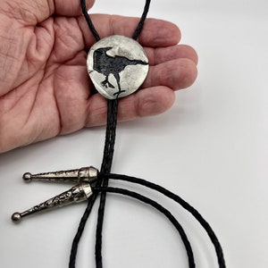 Handmade bolo tie black crow on pewter showing leather braided cord with tips. Artist's hand holding pewter crow.