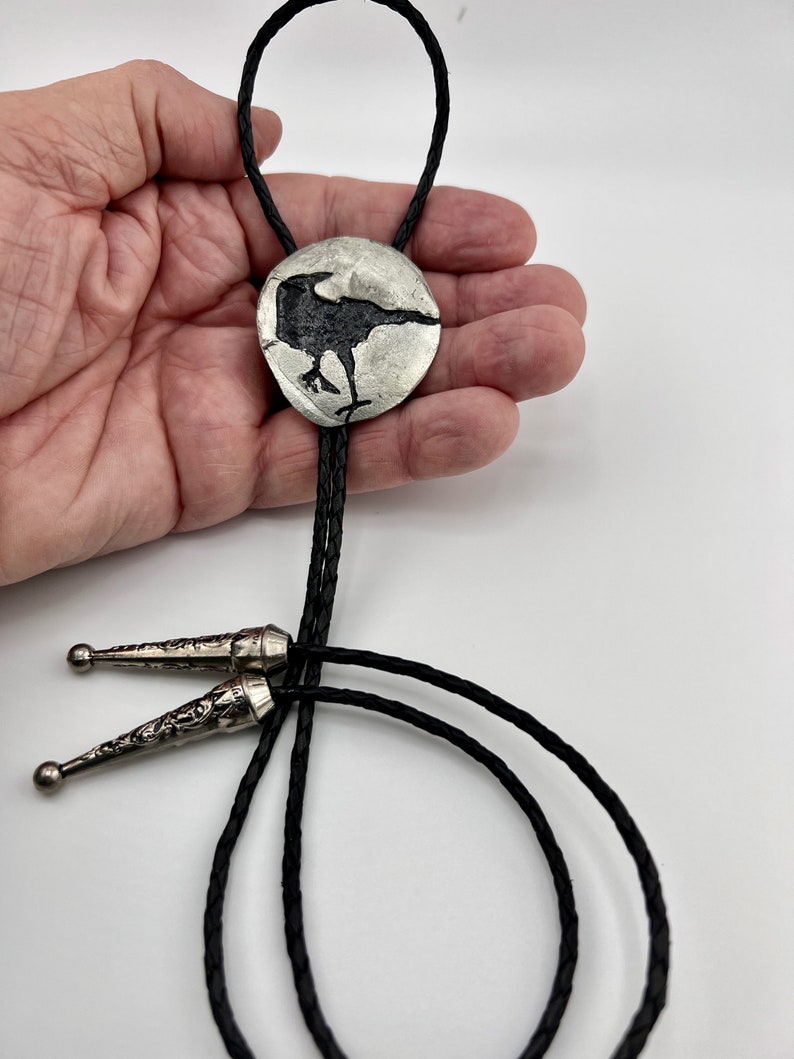 Handmade bolo tie black crow on pewter showing leather braided cord with tips. Artist's hand holding pewter crow.