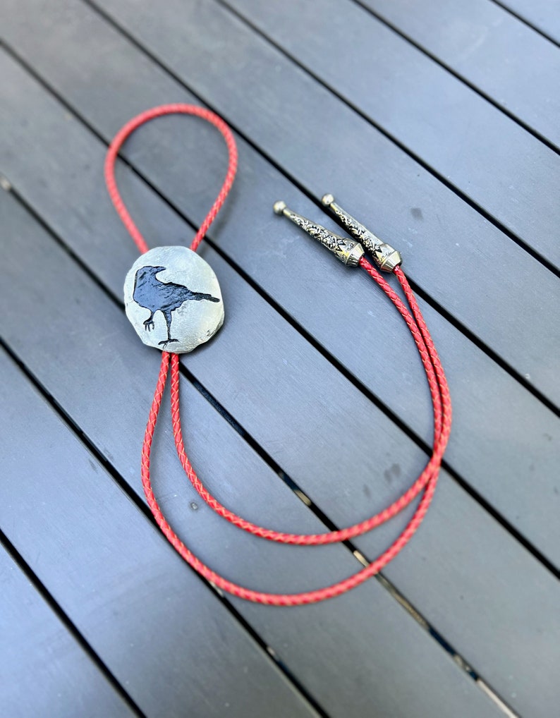 Handmade bolo tie black crow on pewter showing red leather braided cord. Photo taken outside showing the whole bolo tie including decorative tips. Cord length shown is 32 inches but buyer can choose length up to 42 inches.