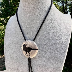 Handmade bolo tie black crow on pewter showing leather braided cord. Crow on gray torso pedestal. Photo taken outside showing shine of the pewter.