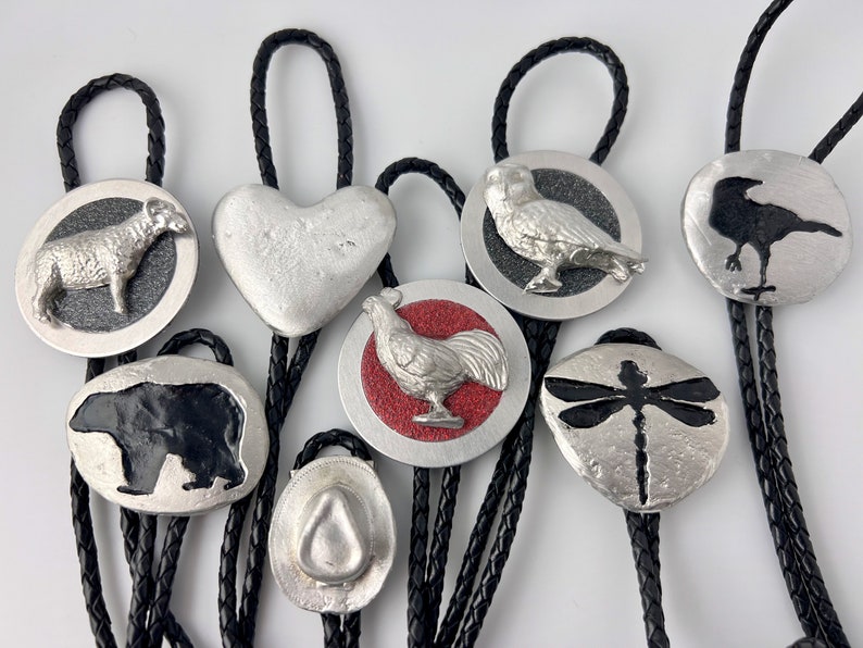 The full collection of pewter-based Bolo Ties from Roads End Productions Co. Each is handmade a Sheep, a Heart, an Owl, a Crow, a Grizzly Bear, a Stetson Hat, a Rooster and a Dragonfly. Each is individually made from casting molten pewter. Legendary.