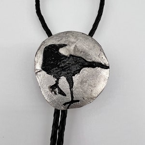 Close-up image of Handmade bolo tie black crow on pewter showing black leather braided cord. The pewter shows the fine detail of the crow captured by the pewter in the silicone mold. A permanent black patina was painted on the crow etching.