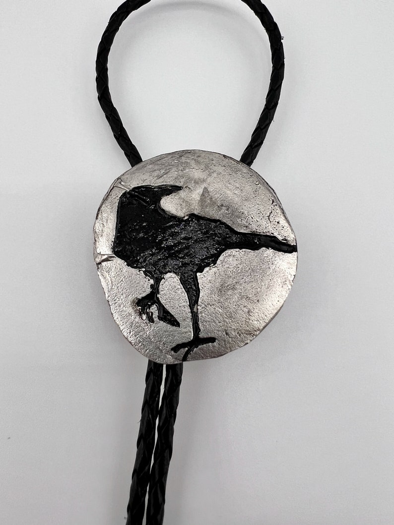 Close-up image of Handmade bolo tie black crow on pewter showing black leather braided cord. The pewter shows the fine detail of the crow captured by the pewter in the silicone mold. A permanent black patina was painted on the crow etching.