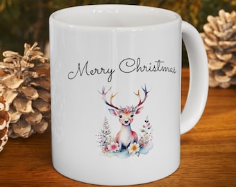 May include: White ceramic mug with a deer illustration and the text "Merry Christmas". The deer has colorful antlers and is surrounded by flowers. The mug is on a wooden surface with pine cones in the background.
