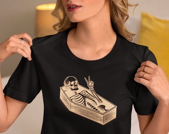 Puede incluir: Camiseta negra con un gráfico de un esqueleto haciendo el signo de la paz mientras está acostado en un ataúd.