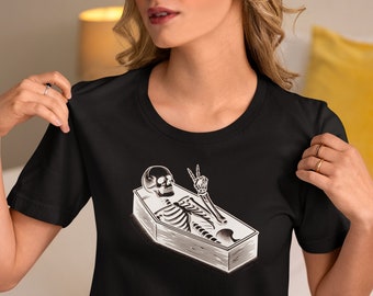 Puede incluir: Camiseta negra con un gráfico de un esqueleto en un ataúd haciendo el signo de la paz.