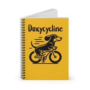 Könnte beinhalten: Ein gelber Spiralblock mit dem Text "Doxycycline" in schwarzen Buchstaben. Unter dem Text befindet sich eine schwarze Silhouette eines Dackels, der Fahrrad fährt.