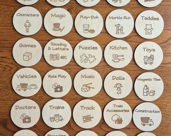 May include: A set of 25 wooden round labels with various images and text. The labels are for organizing children's toys. The labels include categories such as Arts & Crafts, Puzzles & Games, Vehicles, Animals, Books, Characters, Magic, Play-Doh, Marble Run, Teddies, Games, Reading & Letters, Kitchen, Toys, Role Play, Music, Dolls, Magnetic Tiles, Doctors, Trains, Track, Train Accessories, Construction, Bits & Bobs, Dressing Up, Dinosaurs, Balls, and Cars.