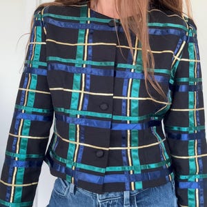 May include: A black long-sleeve jacket featuring a grid pattern of horizontal and vertical ribbons in metallic gold, emerald green, and royal blue. The jacket has a round neckline, long sleeves, and a front button closure with three fabric-covered buttons.