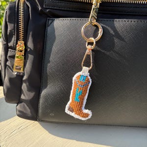 May include: A brown and white stitched cowboy boot keychain with blue accents hangs from a gold zipper. The keychain is attached to a black backpack with gold zippers.
