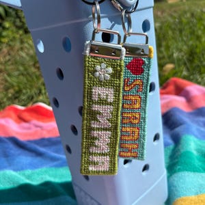 Peut inclure: Deux porte-clés personnalisés brodés avec les noms "EMMA" et "SARAH". Le porte-clés "EMMA" est vert avec une fleur blanche, et le "SARAH" est turquoise avec un cœur rouge. Les deux ont des anneaux argentés.