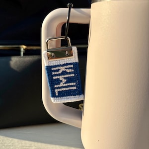 May include: A blue and white embroidered keychain with text is attached to a light pink water bottle. The keychain has a silver clasp and a white border. The water bottle has a white handle.