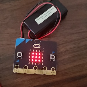 Puede incluir: Una pantalla de matriz LED roja con 9 LED encendidos en una placa de circuito negro. La placa tiene un borde azul y amarillo e incluye dos botones, etiquetados como A y B, y un paquete de baterías con el texto "Solo use baterías de carbono-zinc o alcalinas con su BBC micro:bit".