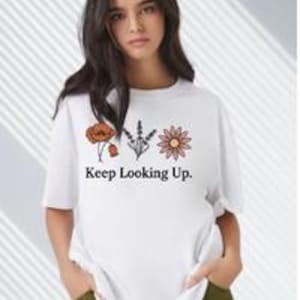 May include: White t-shirt with the text "Keep Looking Up." and three floral illustrations. The flowers are a poppy, lavender, and a daisy. The shirt has short sleeves and a relaxed fit.